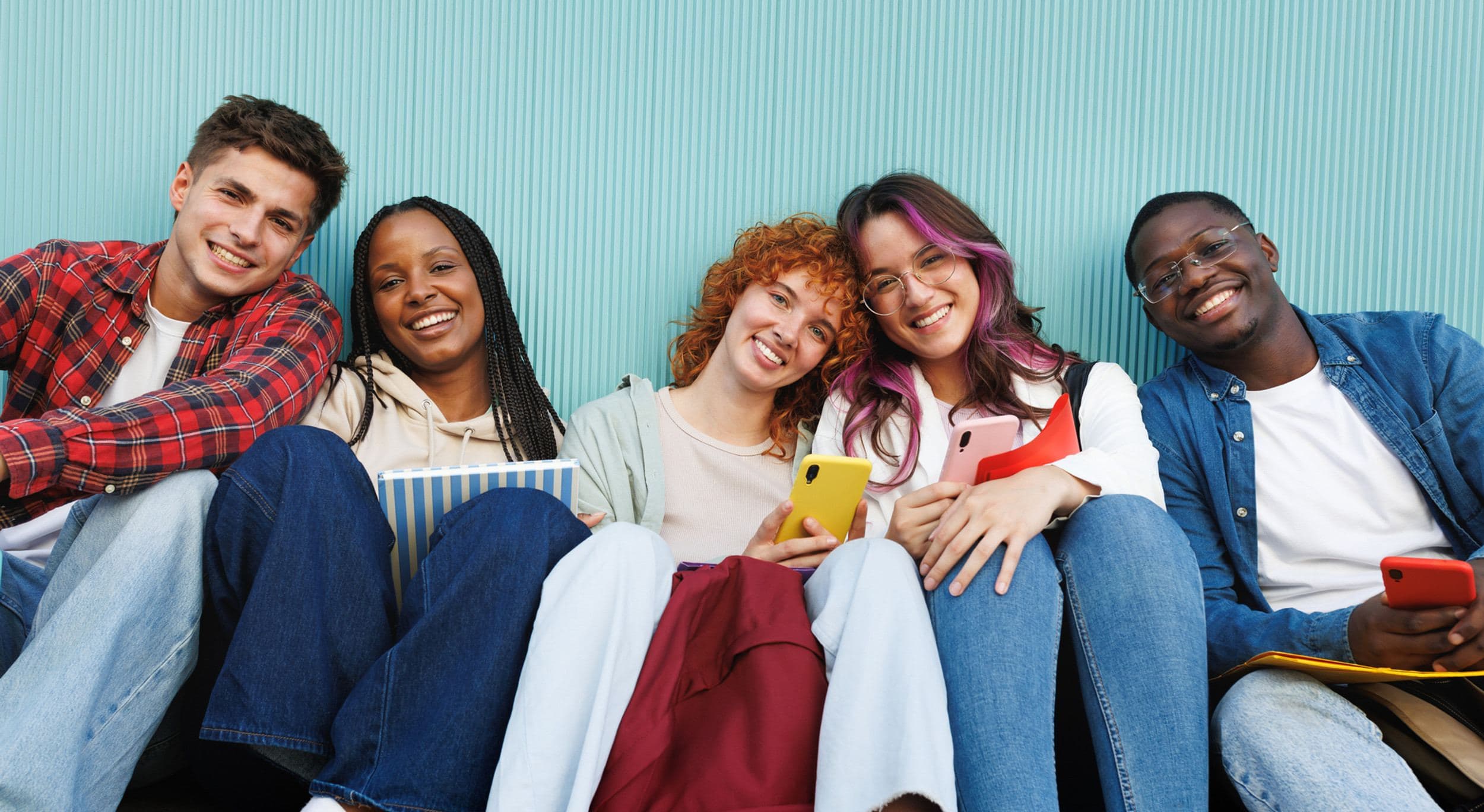 Five young people sitting together against a teal wall, smiling at the camera, some holding phones and notebooks
